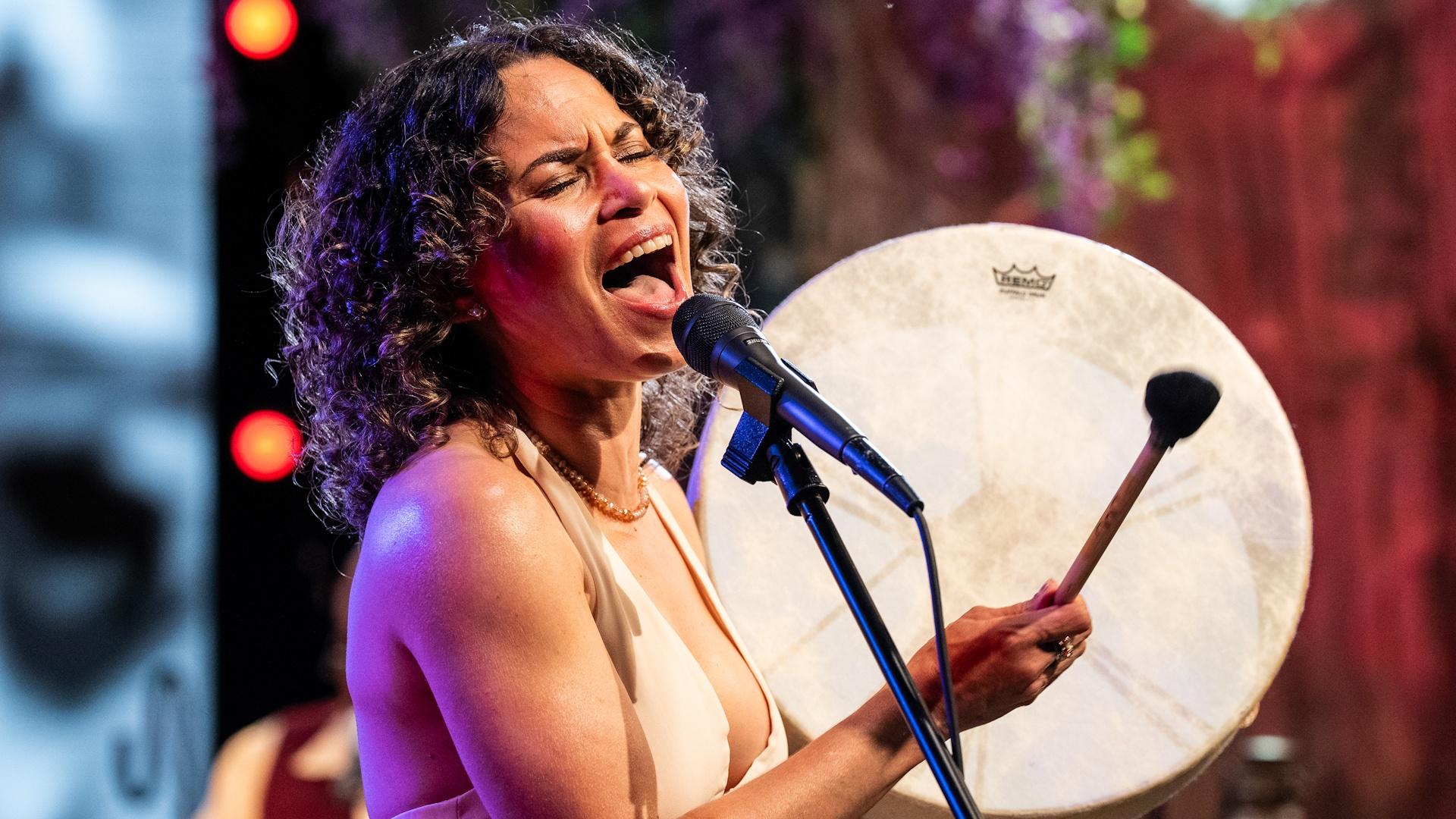 Charly Lowry hitting a handheld drum while singing into a microphone on the set of Shaped by Sound.