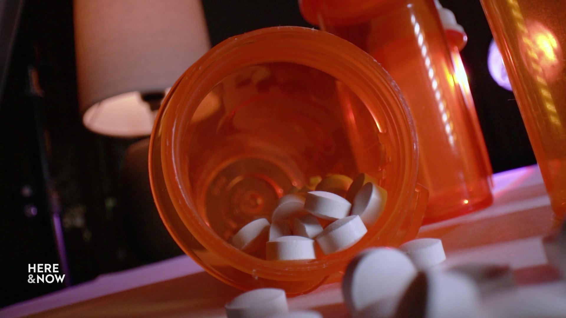 A still image shows a close up of a pill bottle tipped over, with many round white pills spilling out of it.