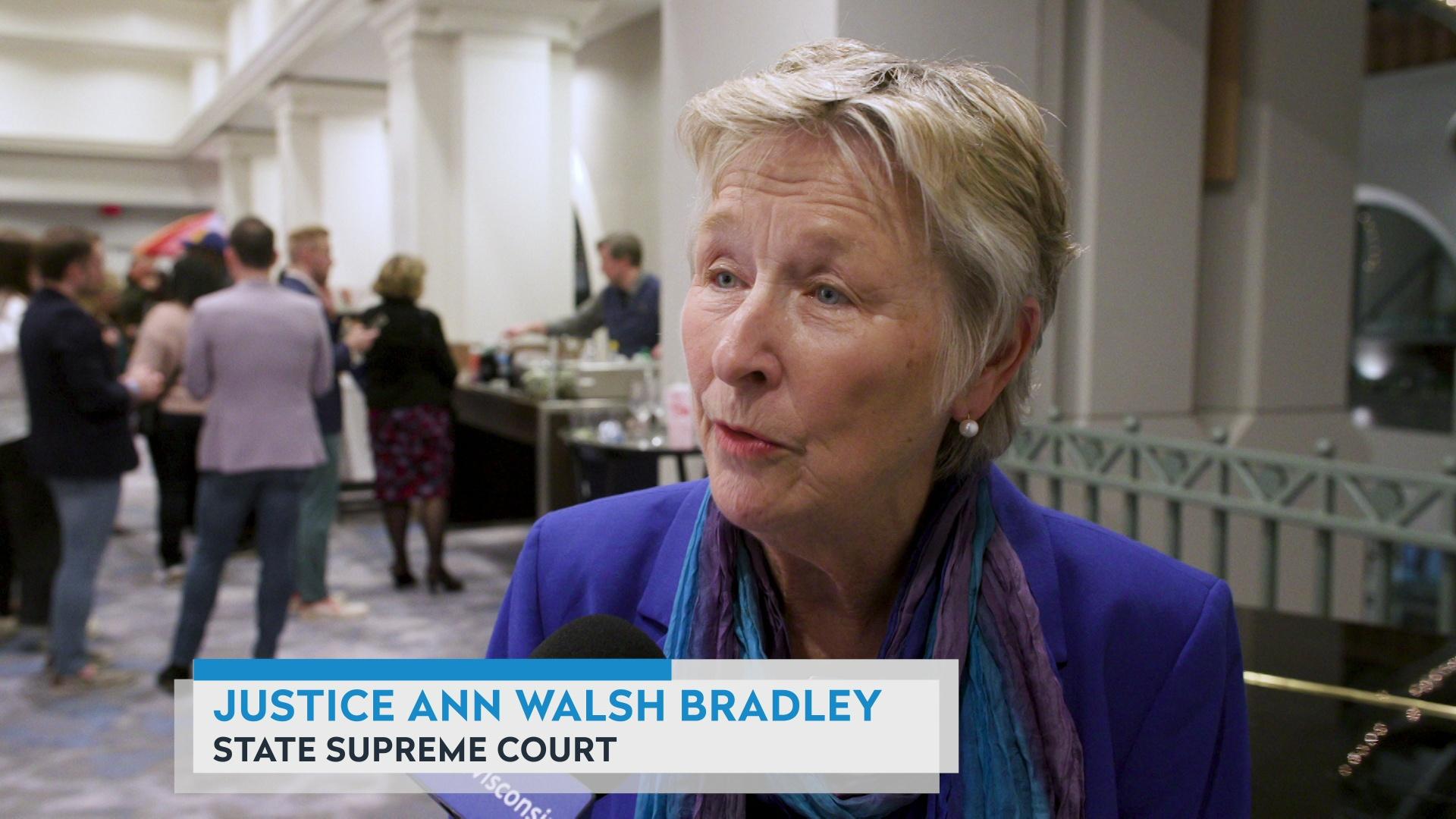 A still image from a video shows Ann Walsh Bradley being interviewed in a large room with people standing in the background and a graphic that reads 'Justice Ann Walsh Bradley' and 'State Supreme Court.'