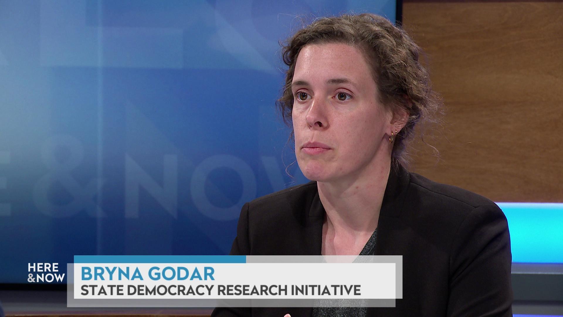 A still image shows Bryna Godar seated at the 'Here & Now' set featuring wood paneling, with a graphic at bottom reading 'Bryna Godar' and 'State Democracy Research Initiative.' A still image shows Bryna Godar seated at the 'Here & Now' set featuring wood paneling, with a graphic at bottom reading 'Bryna Godar' and 'State Democracy Research Initiative.'