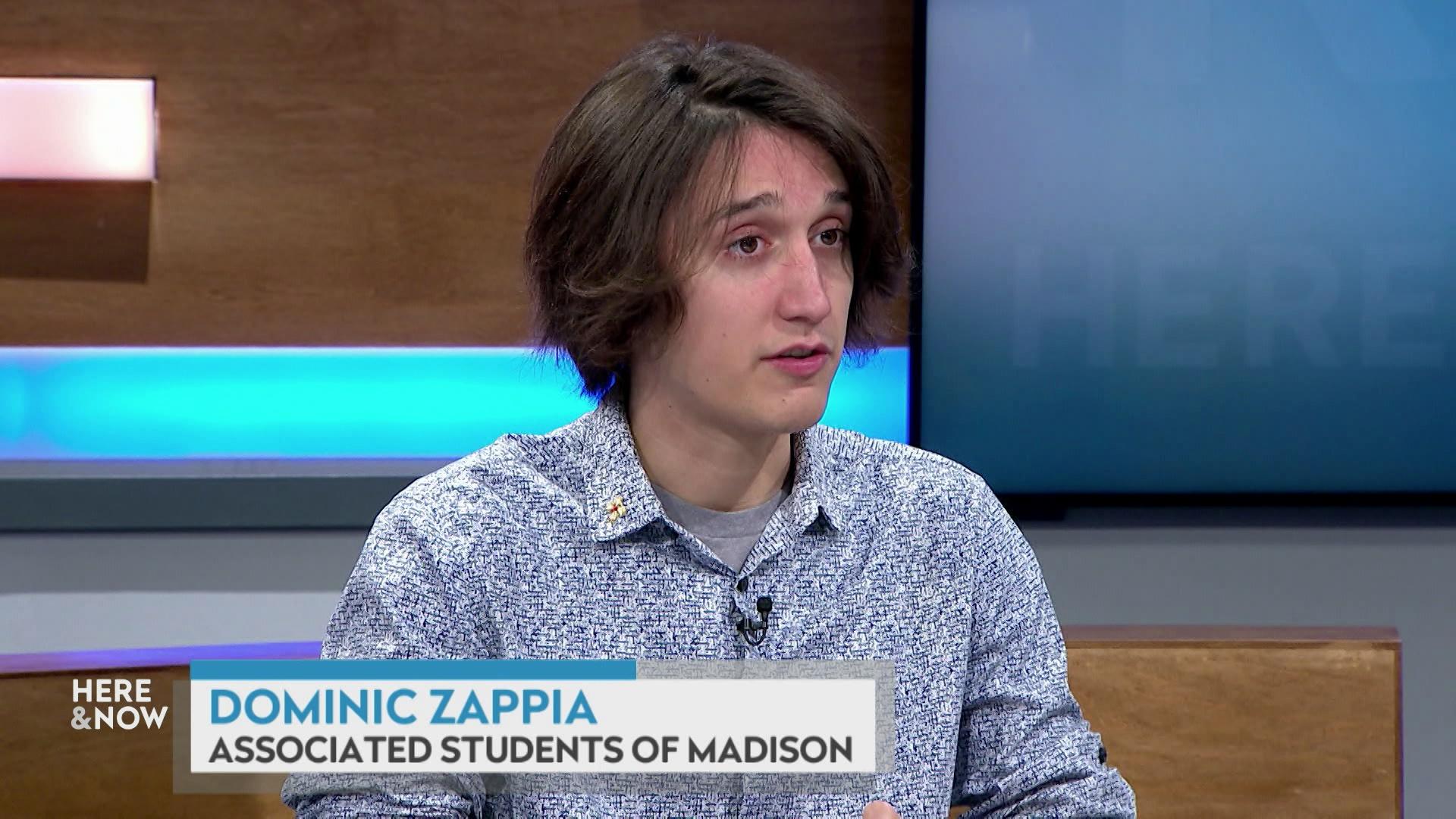 A still image shows Dominic Zappia seated at the 'Here & Now' set featuring wood paneling, with a graphic at bottom reading 'Dominic Zappia' and 'Associated Students of Madison.'