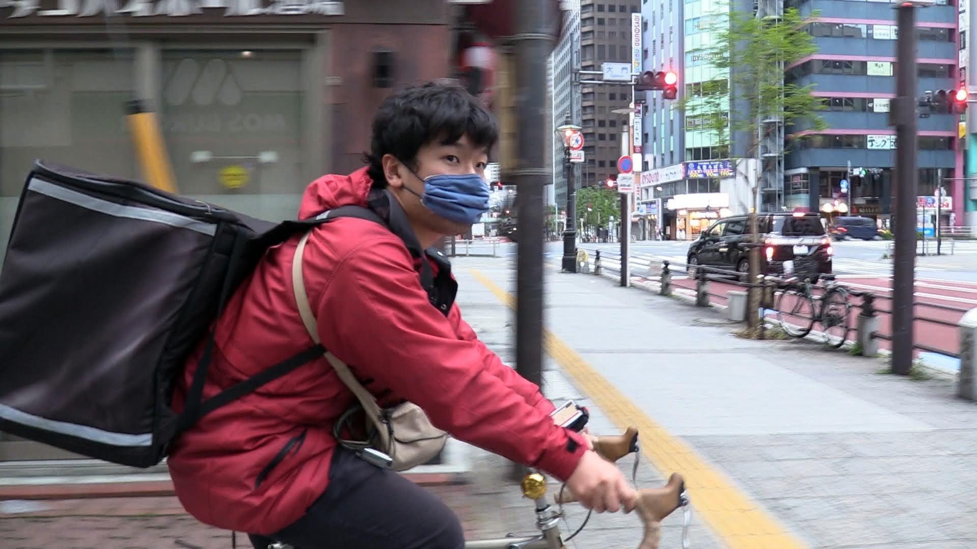 A young man in a red coat on a bike with a large black case strapped to his back