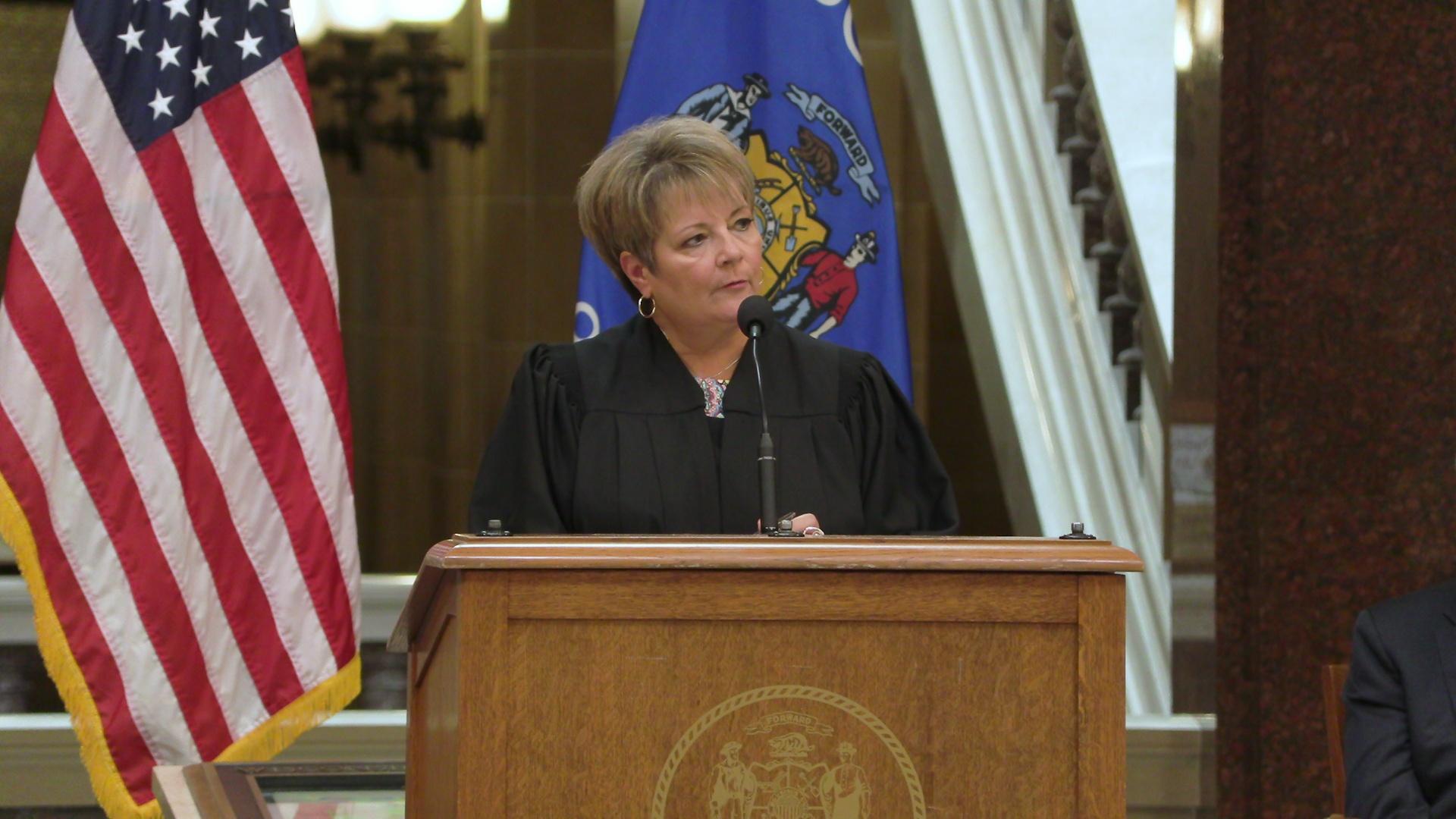 Janet Protasiewitcz dressed in a black gown stands behind a wooden podium and microphone with the U.S. and Wisconsin state flags on each side of her.